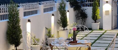 An outdoor seating area with white metal chairs and tables, surrounded by potted plants and a white fence.