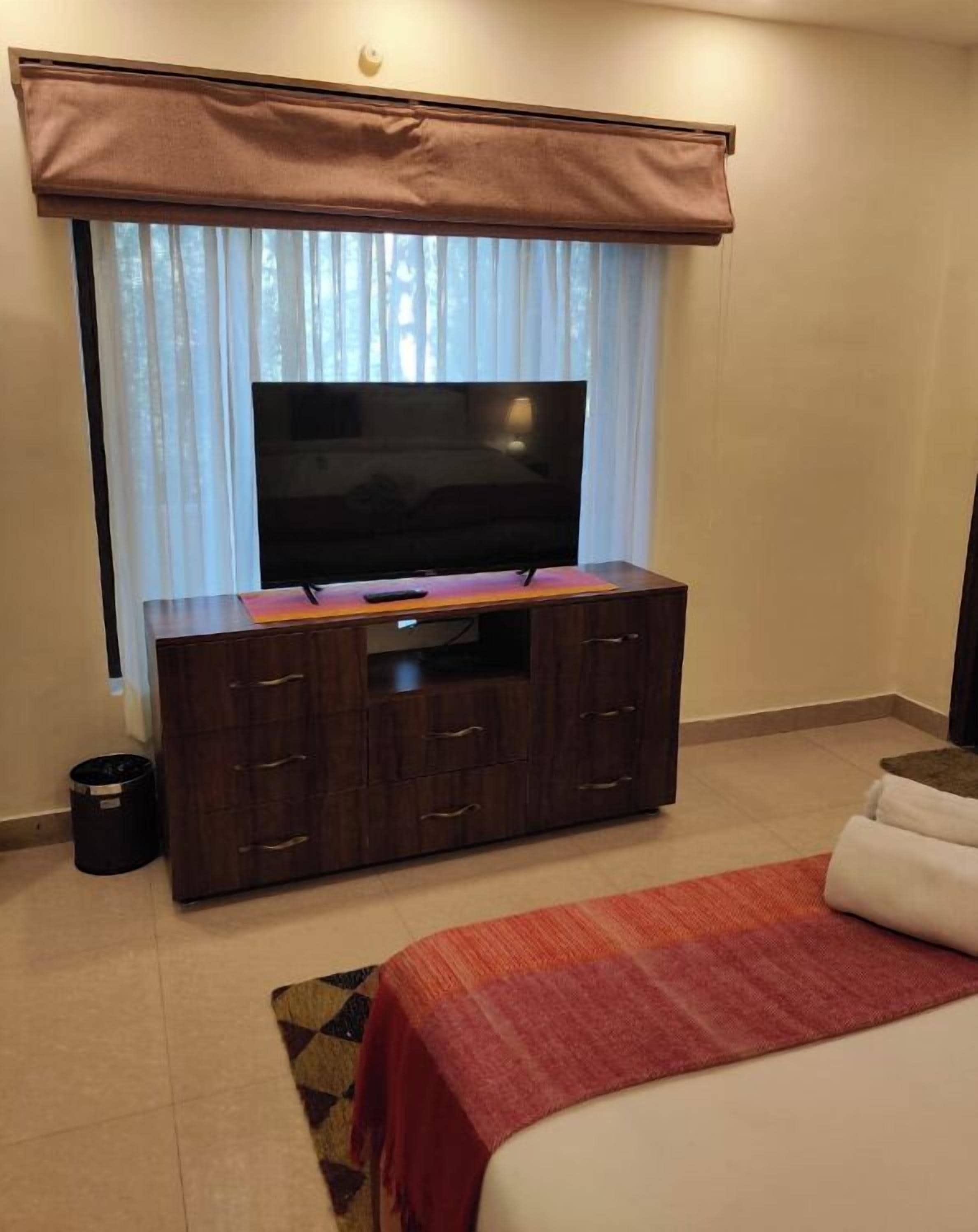 Ein Hotelzimmer mit einem großen Flachbildfernseher auf einer Holzkommode, einem Bett mit roter Decke und einem Fenster mit brauner Gardine.