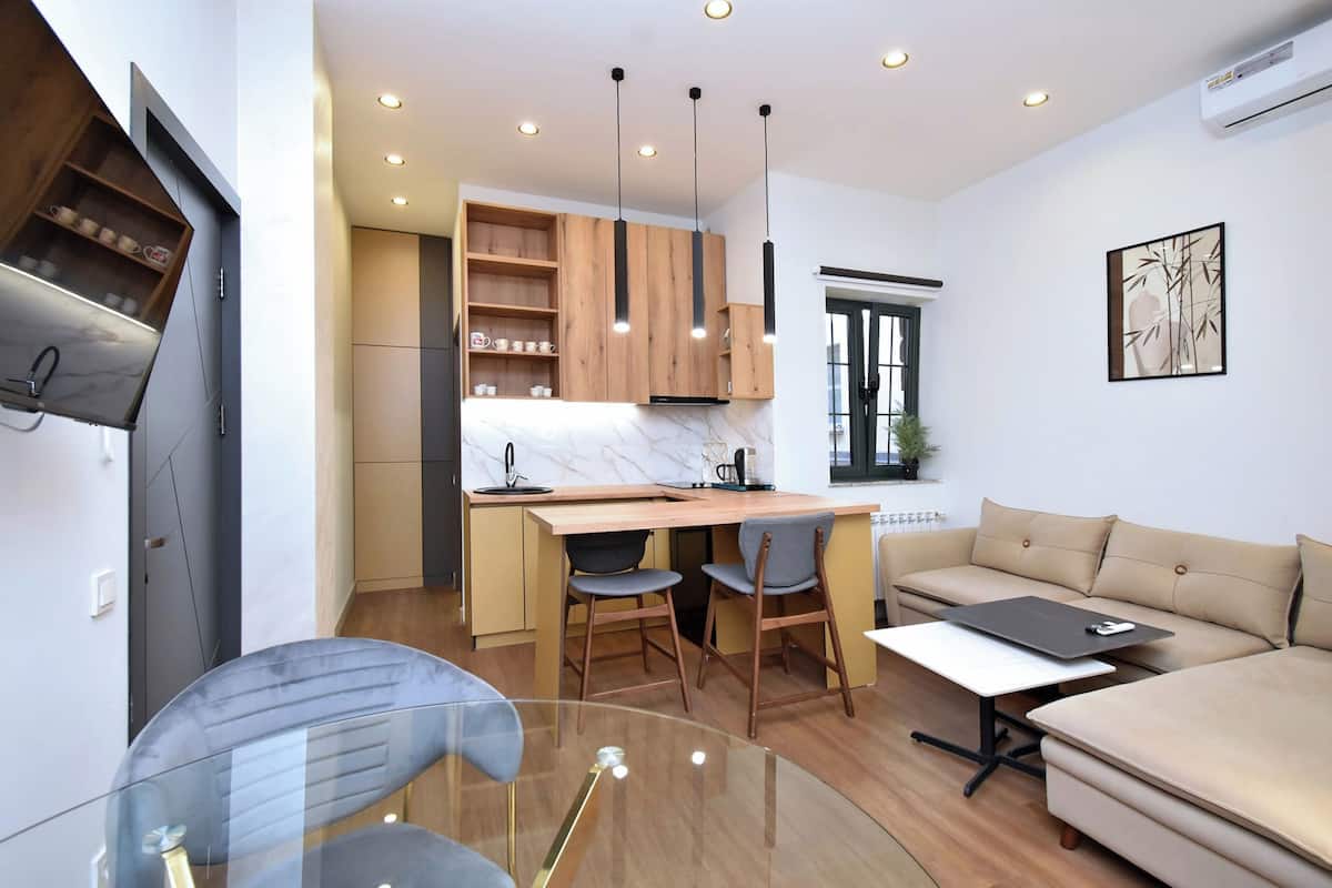 A modern kitchen with wooden cabinets, a built-in island, and pendant lighting. There is a dining area with a glass table and chairs, and a sofa in the living room.