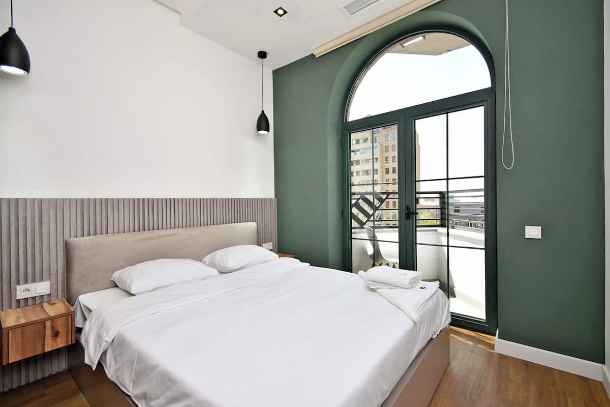 A modern bedroom with a large bed, a wooden nightstand, a black pendant light, and a view of the city through a large arched window.
