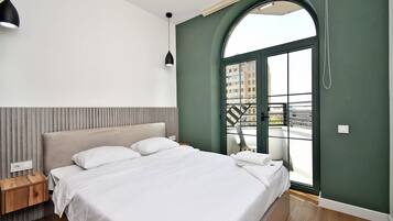 A modern bedroom with a large bed, a wooden nightstand, a black pendant light, and a view of the city through a large arched window.