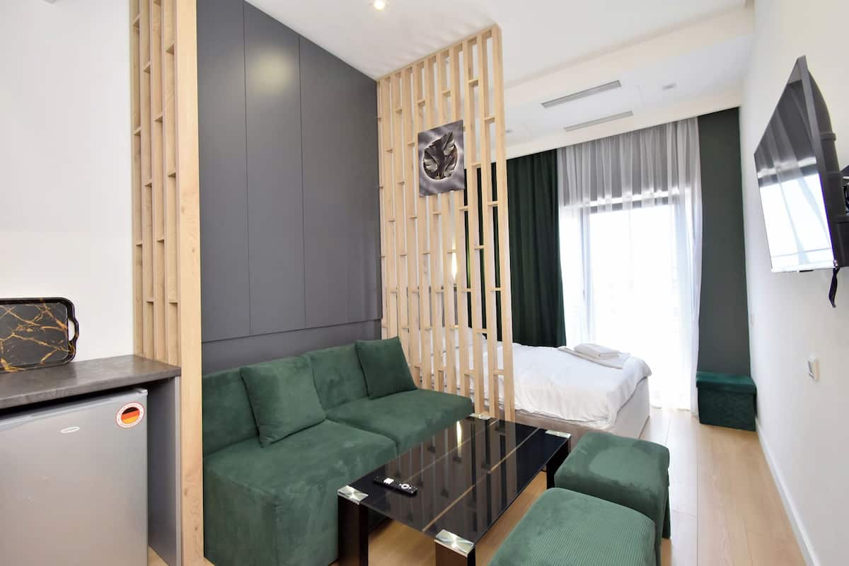 A modern hotel room with a green sofa, a small table, a bed with white linens, a wooden partition, and a large window with curtains.
