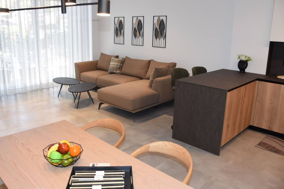 Ein modernes Wohnzimmer mit einer Couch, einem Esstisch mit Stühlen, einer Obstschale und einer kleinen Küchenzeile mit einer Arbeitsplatte und Barhocker.