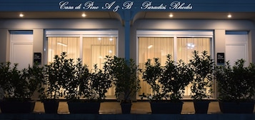 Fachada de hotel com duas janelas grandes, plantas em vasos na frente e uma placa acima das janelas.
