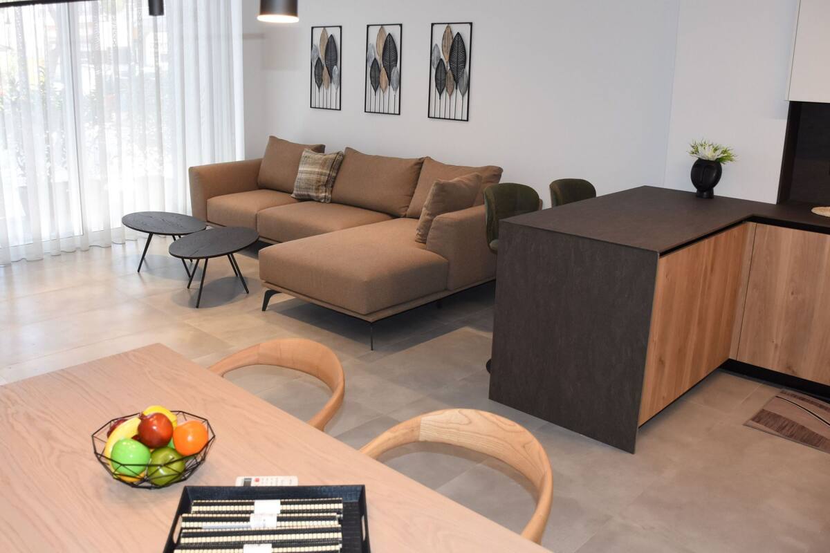 Een moderne woonkamer met een bank, een eettafel met stoelen, een fruitschaal en een klein keukentje met een aanrecht en krukken.