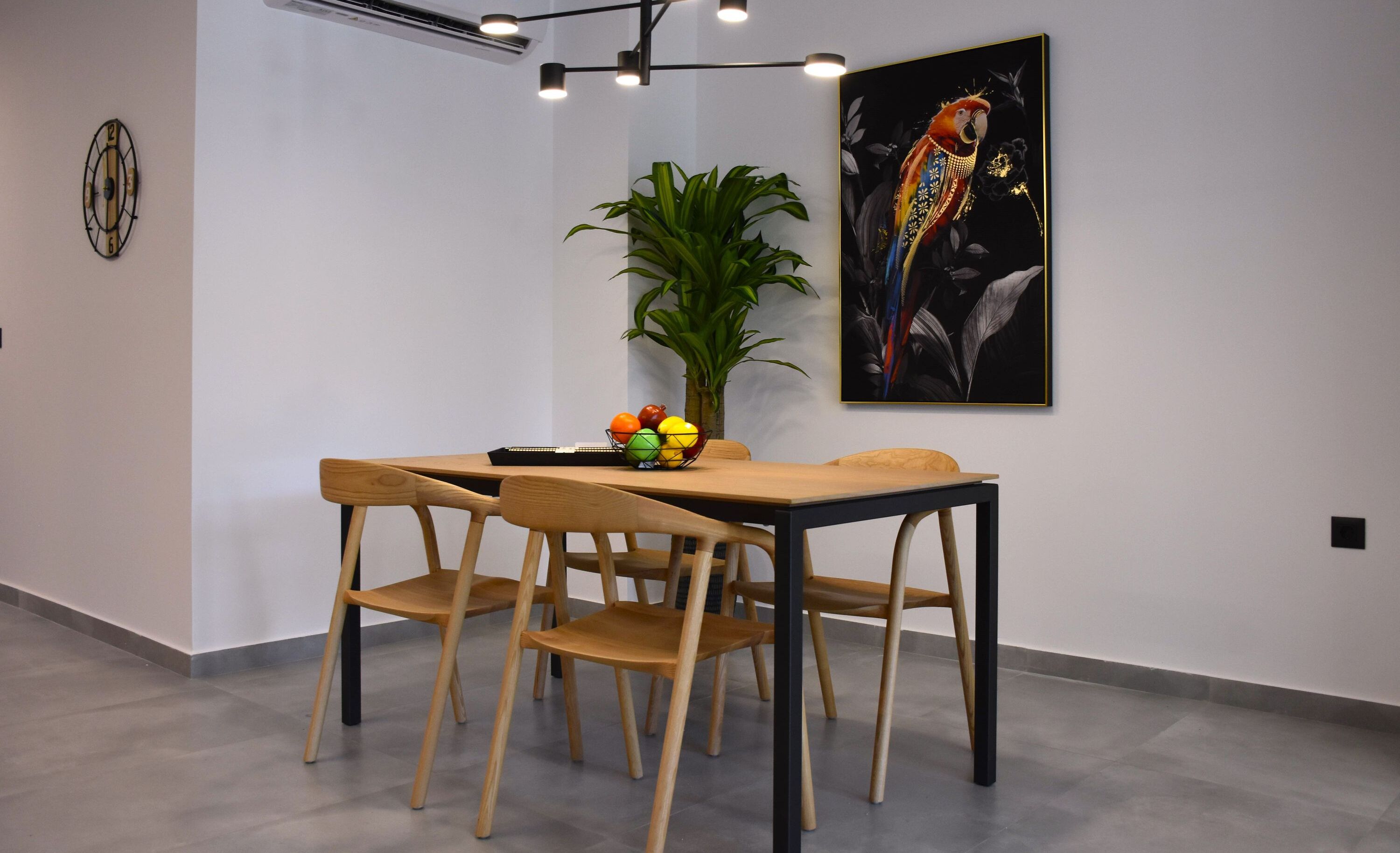 A dining area with a wooden table and chairs, a fruit bowl, a potted plant, and a framed artwork on the wall.