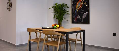 A dining area with a wooden table and chairs, a fruit bowl, a potted plant, and a framed artwork on the wall.