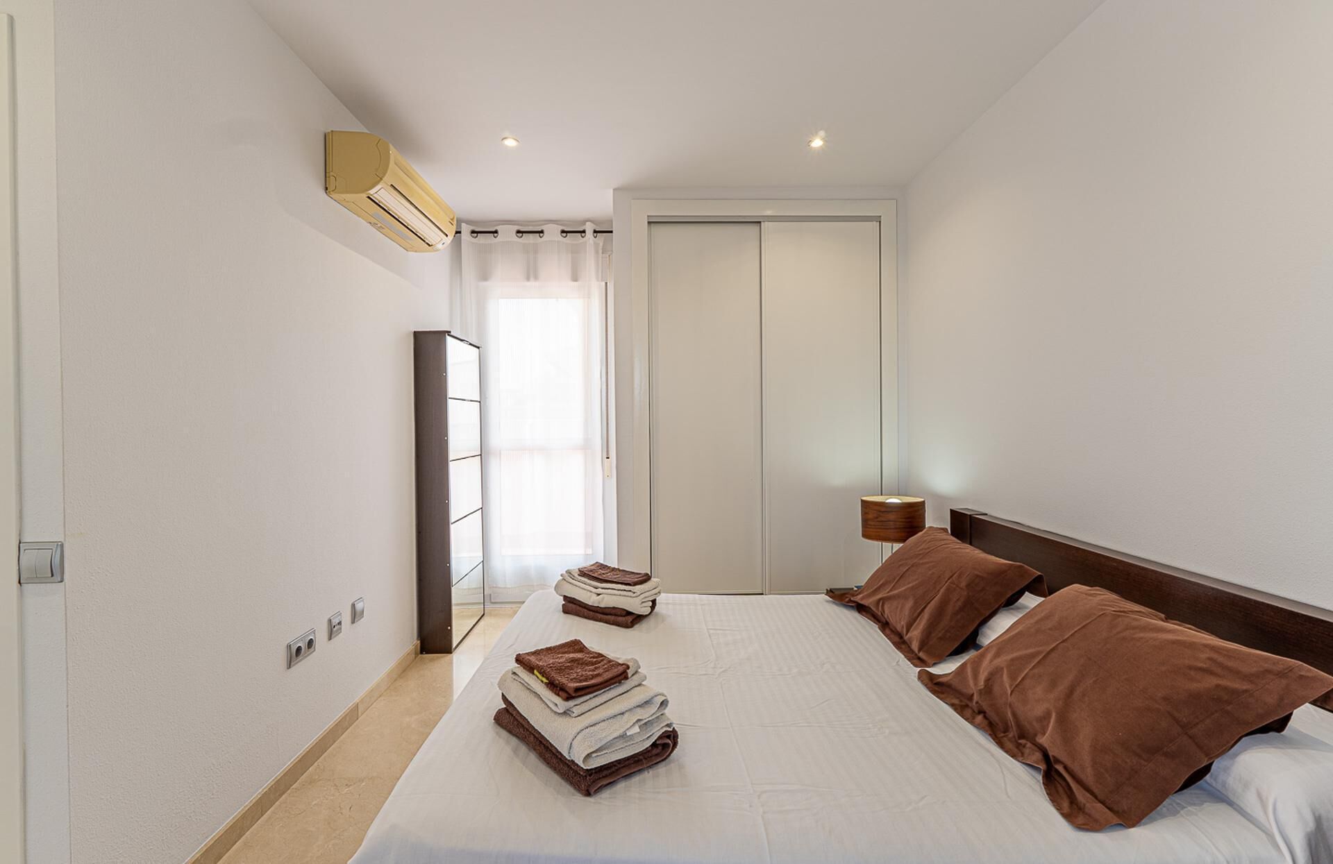 A hotel room with a bed, a bedside lamp, a wardrobe, and an air conditioning unit.