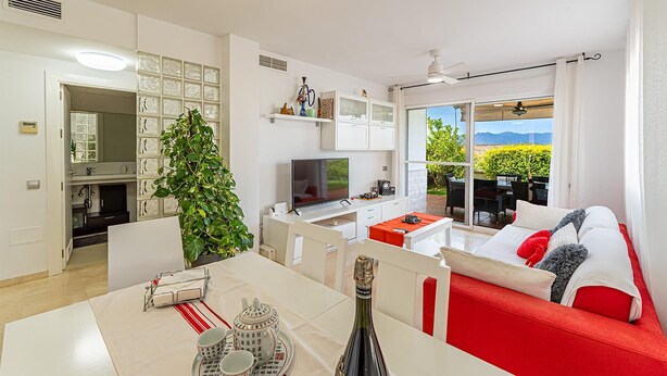 Un salon moderne avec un téléviseur, un coin repas avec une table préparée pour le thé, et un balcon offrant une vue sur l’extérieur.