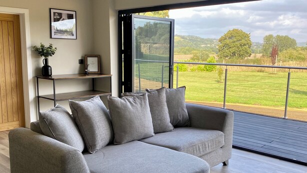 A modern living room with a grey sectional sofa, a wooden floor, a glass door leading to a balcony, and a framed picture on the wall. - The Orchard Retreat  (Corston)