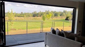 A modern living room with a grey sofa, a wooden floor, and a large glass door leading to an outdoor area with a metal railing and a view of a green field. - The Orchard Retreat  (Corston)