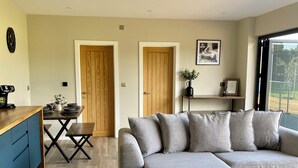 A modern living room with a grey sectional sofa, wooden dining table, and a kitchen area with a blue cabinet. - The Orchard Retreat  (Corston)