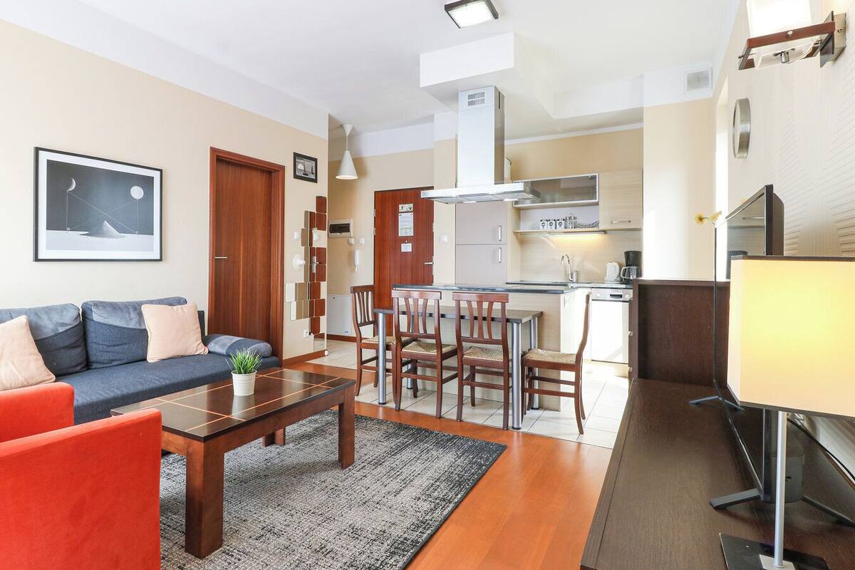 A modern living room with a sofa, coffee table, and chairs. A kitchen with a stove and sink is visible in the background.