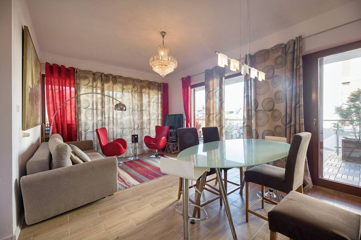 A modern living room with a glass dining table, red chairs, and a chandelier.