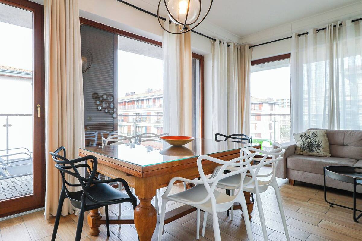 A modern living room with a wooden dining table, white and black chairs, a glass-top table, a sofa, and a balcony with a view of buildings.