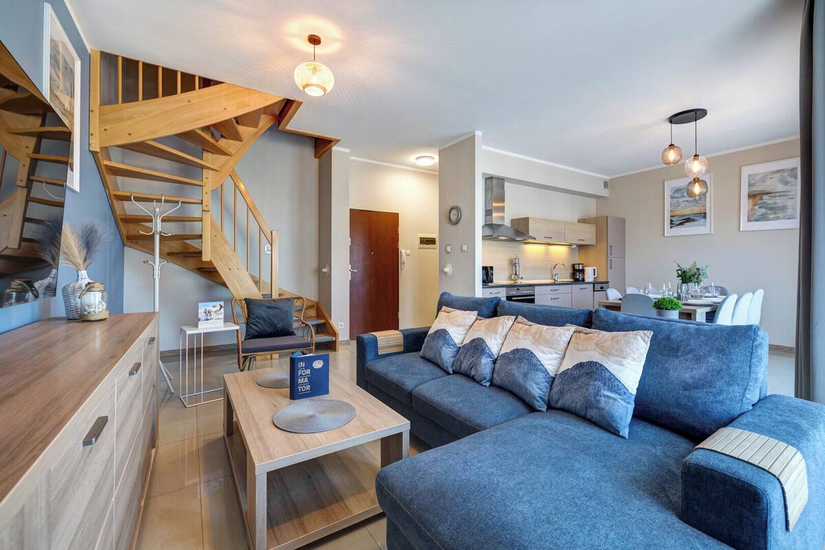 A modern living room with a blue sofa, wooden staircase, and a kitchen in the background.
