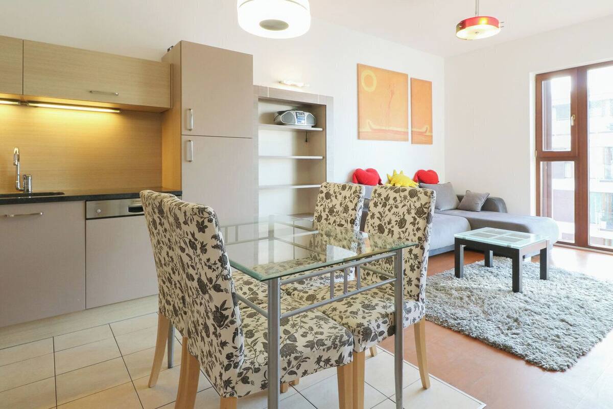 A modern kitchen with a dining area, featuring a glass table and floral chairs, a sofa, and a large window.