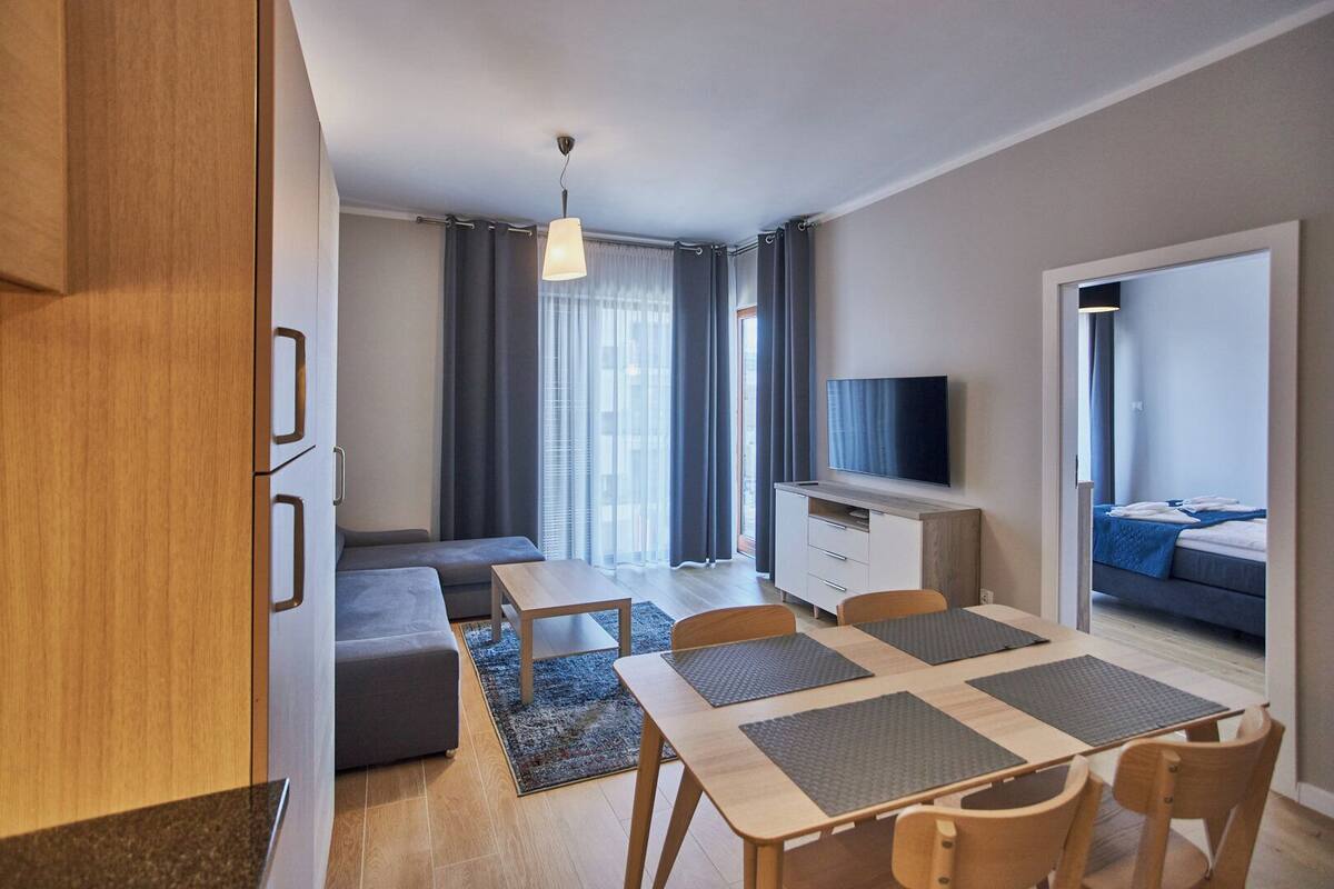 A modern living room with a sofa, a dining table with chairs, a TV, and a view of a bedroom through an open door.