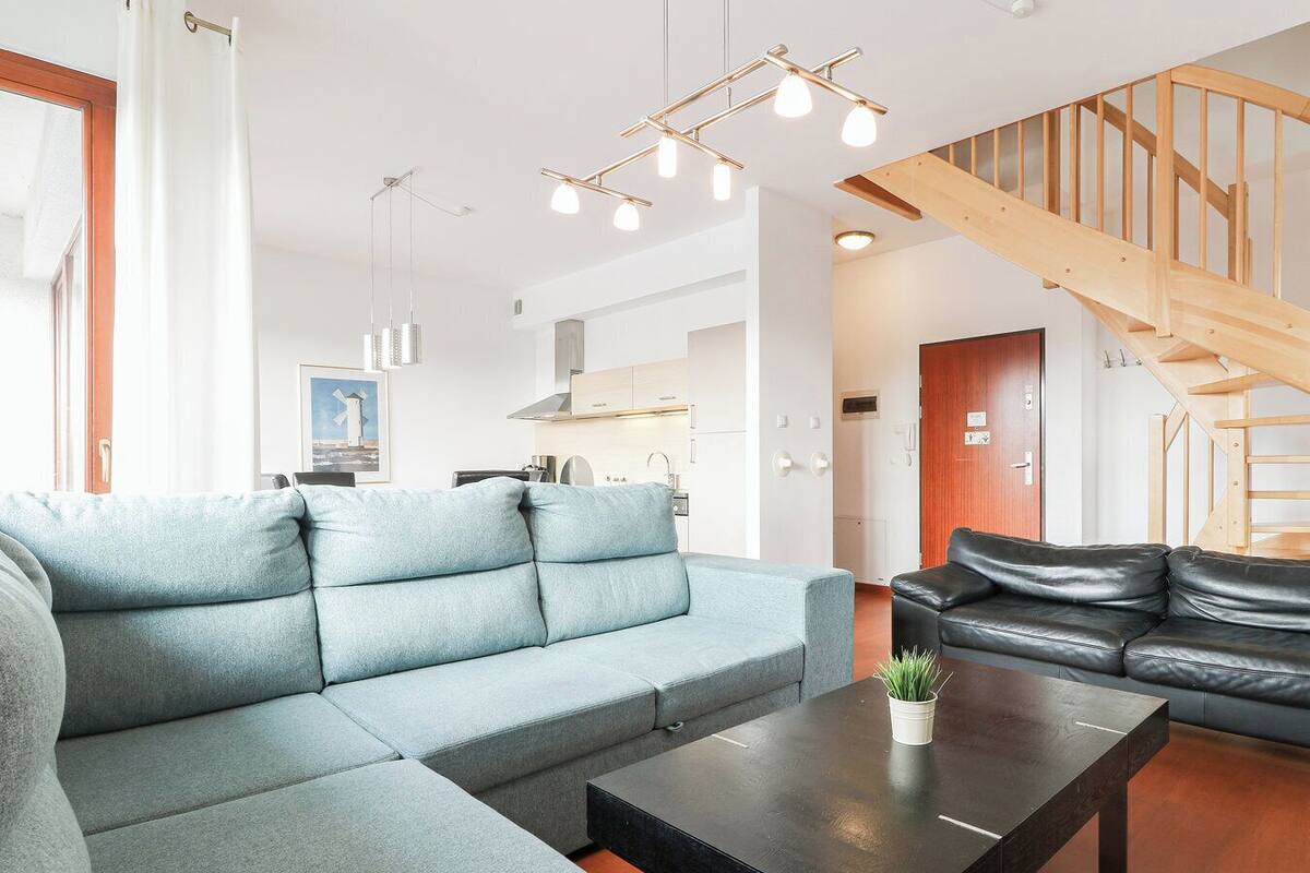 A modern living room with a sectional sofa, a coffee table, and a black leather sofa. A staircase with wooden railings is visible in the background.