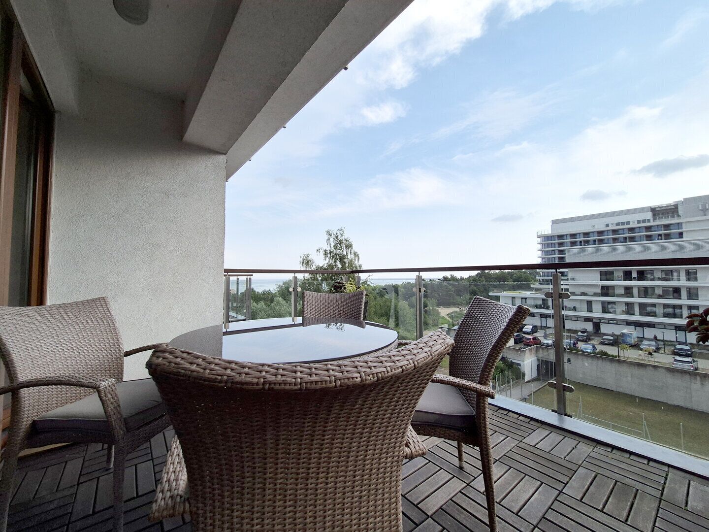 A balcony with wicker furniture, a glass table, and a view of a modern building and greenery.