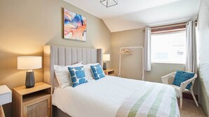 Une chambre d’hôtel bien aménagée, avec un grand lit, des lampes de chevet, une petite table d’appoint, une œuvre d’art accrochée au mur, une chaise et une fenêtre avec des rideaux.