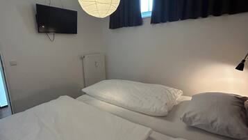 A bedroom with two beds, a flat-screen TV mounted on the wall, a ceiling light fixture, and a window with dark curtains.