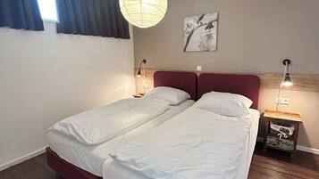 A double bed with white linens, a red headboard, a hanging light fixture, a nightstand with a lamp, and a wall-mounted picture.