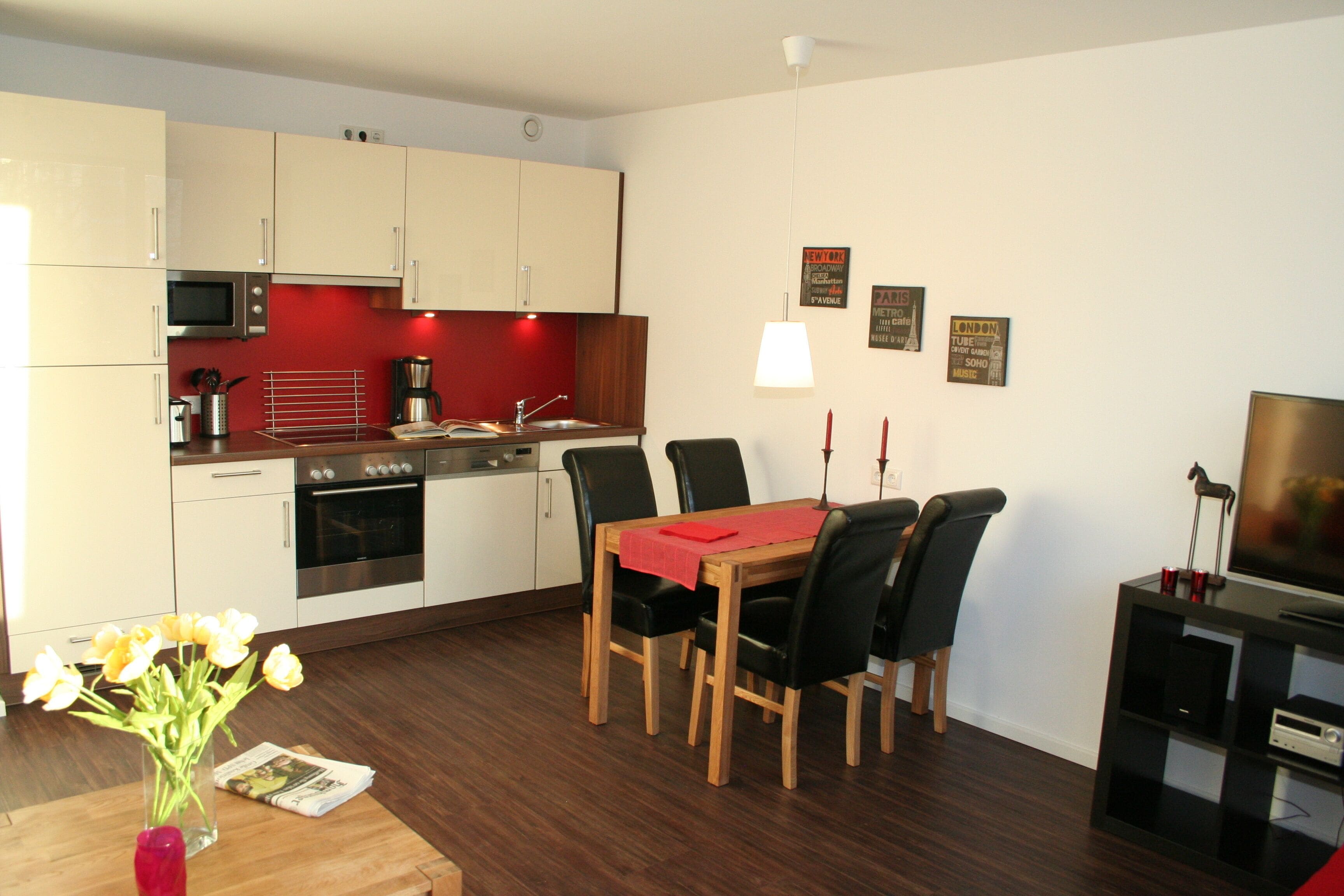 A modern kitchen with white cabinets, a microwave, and a red backsplash. There is a dining table with chairs and a vase of flowers.
