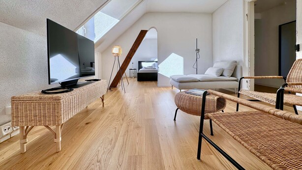 Ein modernes Schlafzimmer im Loft-Stil mit großem Oberlicht, Holzboden, Flachbildfernseher, Rattanmöbeln und einem Bett mit Kopfteil.