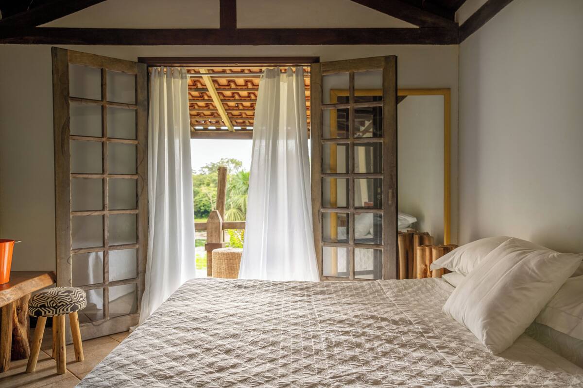 Un dormitorio con cama, un taburete de madera, una mesita y una ventana con cortinas.