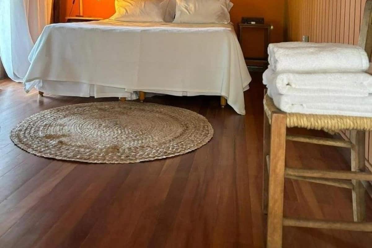 Un dormitorio con cama, lámpara de noche, alfombra redonda tejida, silla con toallas y pared decorada con un cuadro y un objeto decorativo.