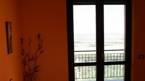 Ein Zimmer mit einem Bett, einem Nachttisch, einer Vase und Blick auf einen Balkon mit Geländer.