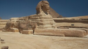 A large stone structure resembling a pyramid with a distinct headpiece, surrounded by other similar stone blocks and a clear blue sky. - Pyramids Kingdom  (Giza)