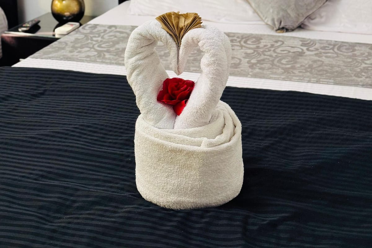 A bed with a black headboard, white pillows, and a decorative towel shaped like a swan holding a red rose.