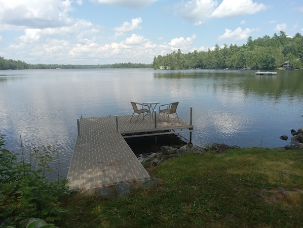 A dock with two chairs and a table, overlooking a calm lake with a forested shoreline.