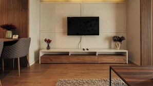 A modern living room with a flat-screen TV mounted on a white wall, wooden flooring, a grey chair, a small white console table with decorative items, and a wooden coffee table. - Nest Inn Magic Apartment Scala Center (Cluj-Napoca)