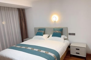 Una cama bien tendida con una colcha estampada, dos almohadas, una mesita de noche con cajones y una lámpara fijada a la pared.