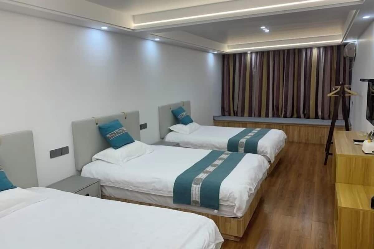 A hotel room with three beds, a wooden floor, a window with striped curtains, and a wooden desk with a lamp.
