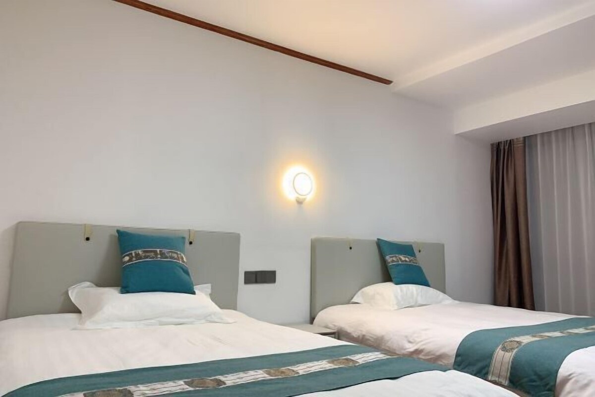 A hotel room with two beds, each with a headboard, white bedding, teal throw pillows, and a teal throw blanket. There is a wall-mounted light fixture above the beds.