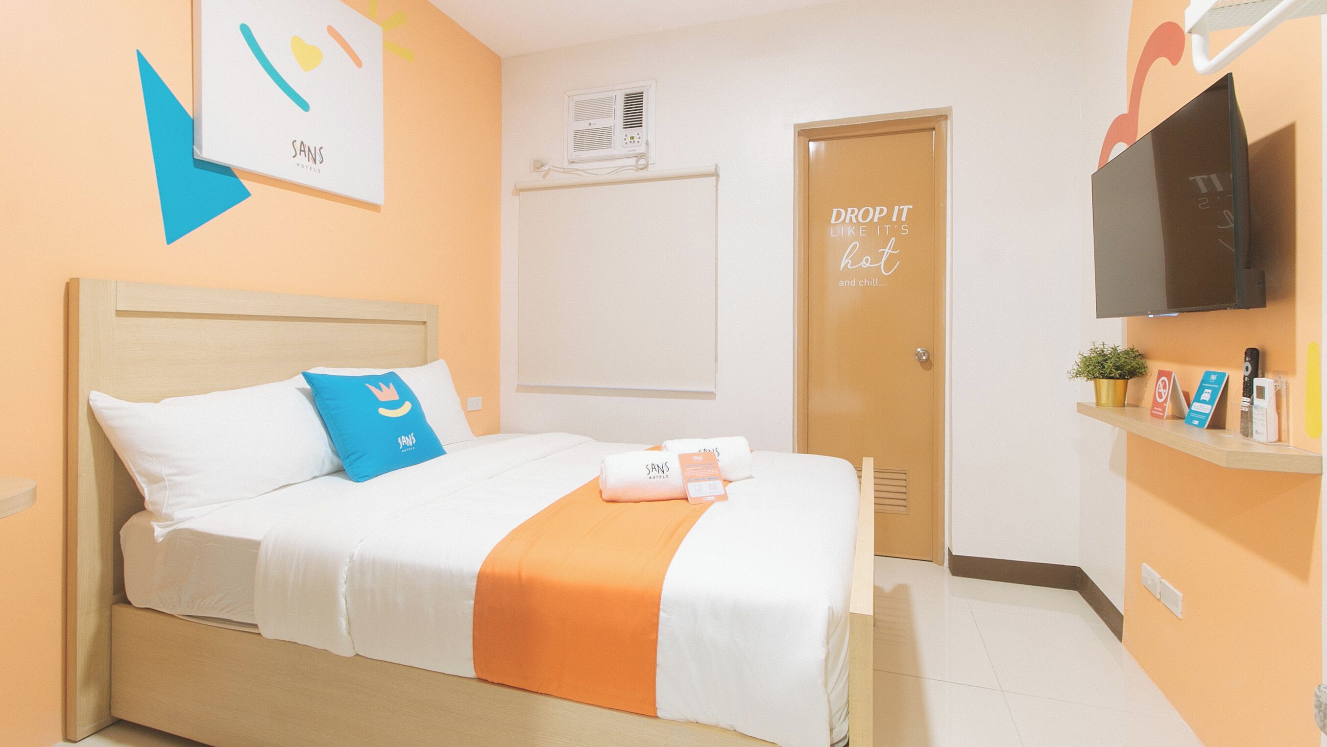 A hotel room with a bed, a TV, a door with a sign, and a wall-mounted air conditioner.