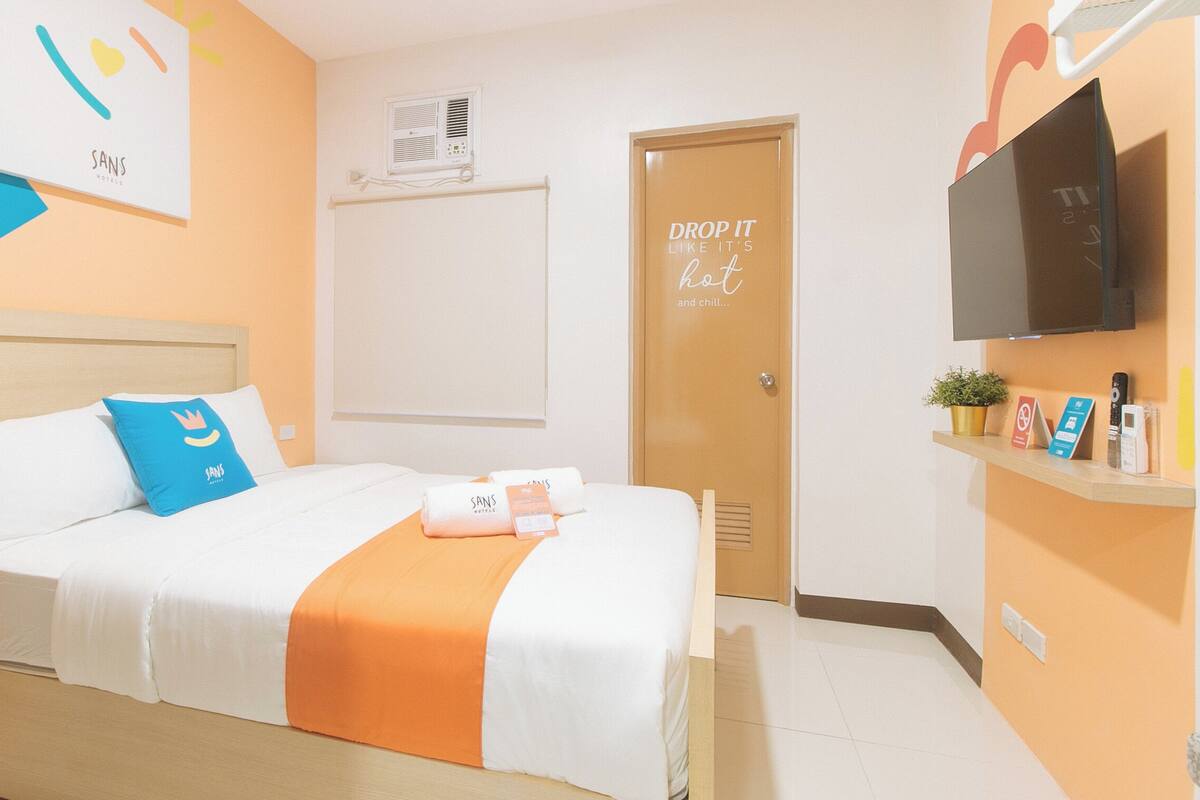 A hotel room with a bed, a TV, a door with a sign, and a wall-mounted air conditioner.