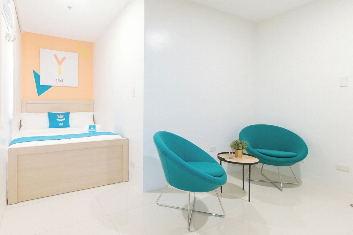 Une chambre d’hôtel avec un lit, une œuvre d’art accrochée au mur, une petite table avec une plante et deux fauteuils de couleur turquoise.