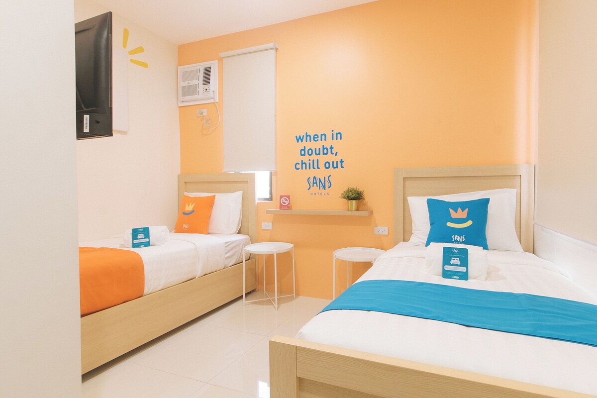 Habitación de hotel con dos camas, una mesita, aire acondicionado instalado en la pared y un cartel con el nombre del hotel.