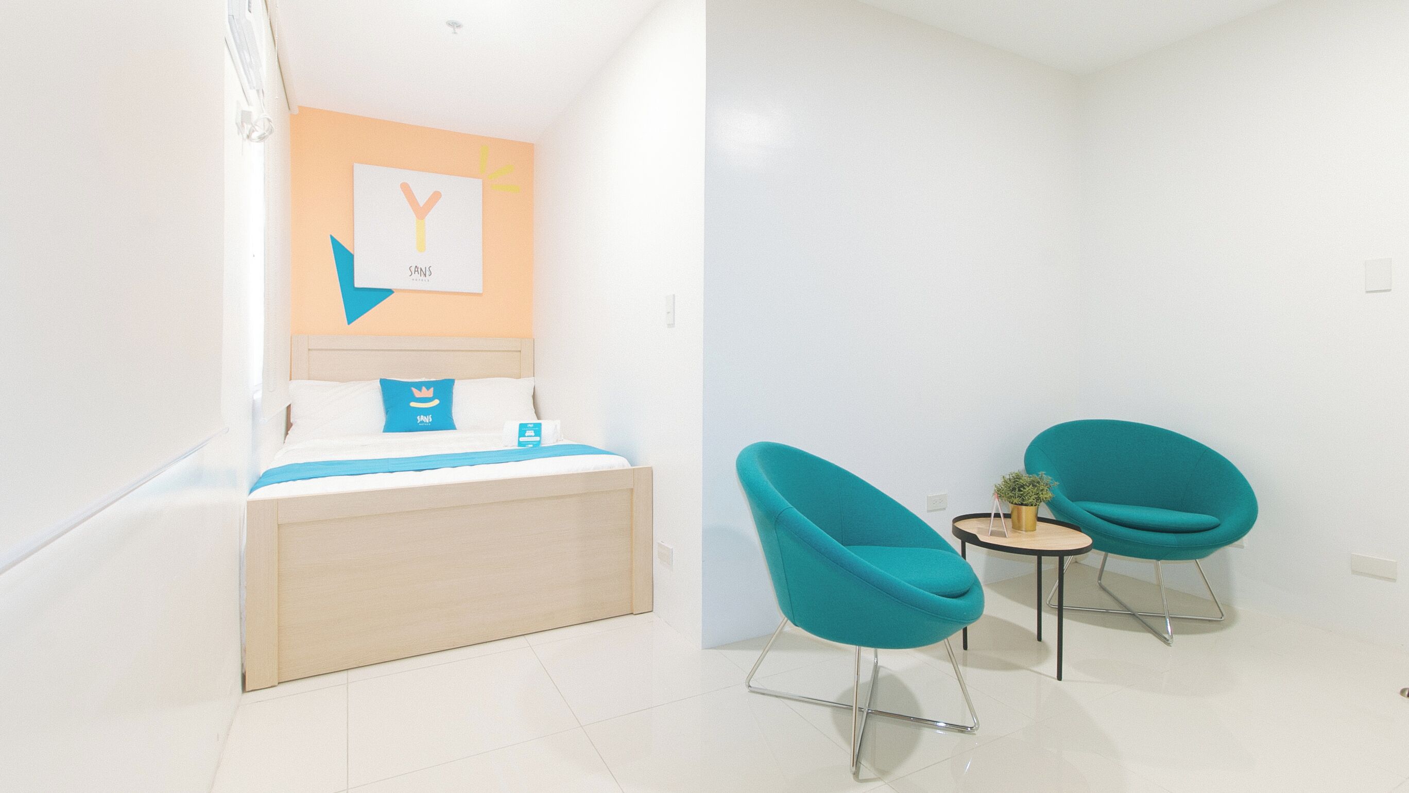 A hotel room with a bed, a wall-mounted artwork, a small table with a plant, and two teal chairs.