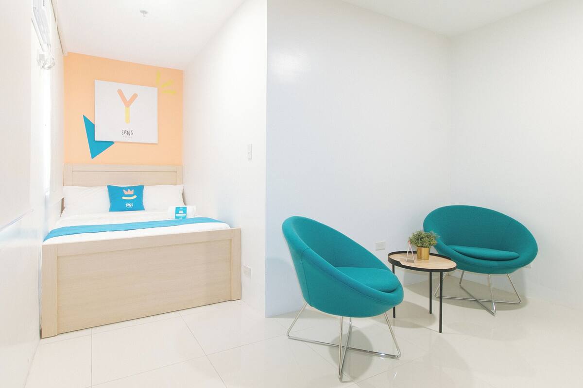 A hotel room with a bed, a wall-mounted artwork, a small table with a plant, and two teal chairs.