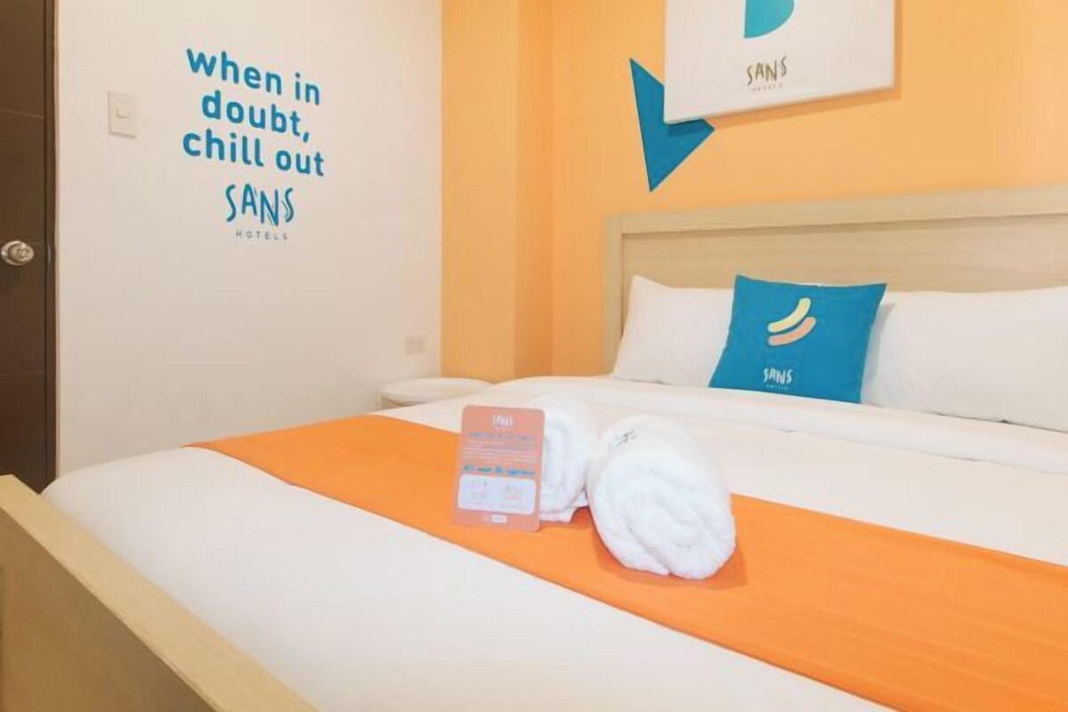 Une chambre d’hôtel avec un lit, un oreiller bleu, une serviette et un panneau mural sur lequel on peut lire : « En cas de doute, détendez-vous, SANS HOTELS ».