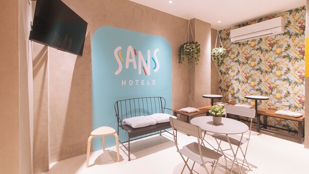 Una habitación de hotel moderna con una pared azul que presenta el nombre 'Sans Hotels'. Hay una cama con estructura metálica, un taburete pequeño, una mesa redonda con una planta en maceta y una planta colgante.