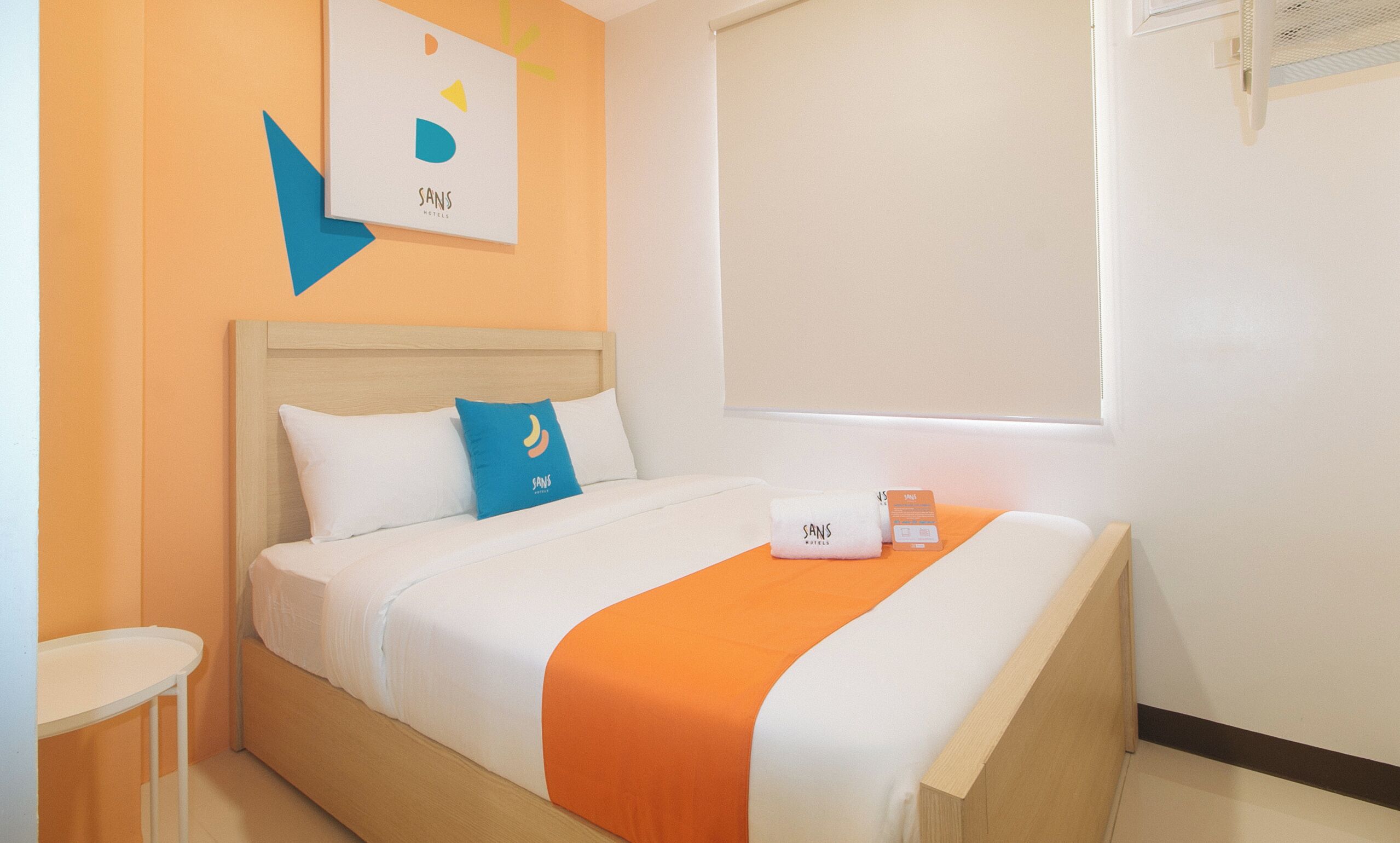 A hotel room with a bed, a bedside table, a window with blinds, and a wall-mounted air conditioning unit.