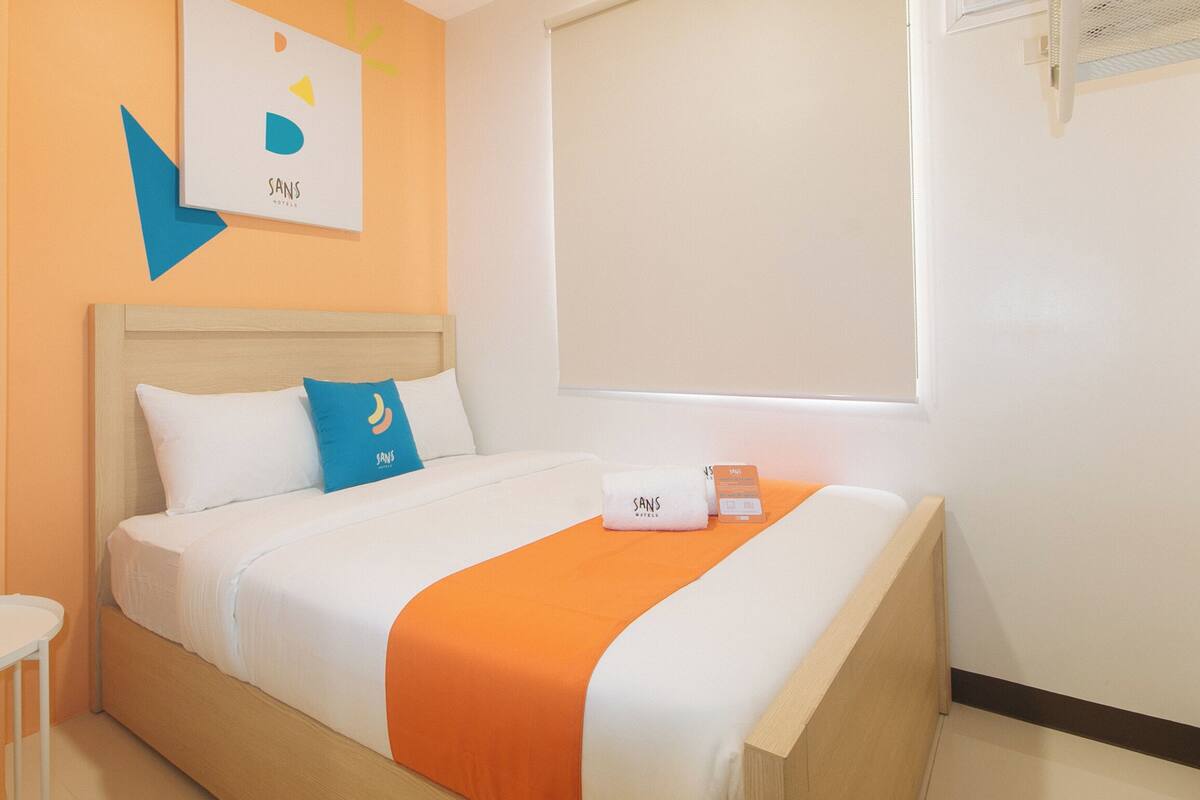 A hotel room with a bed, a bedside table, a window with blinds, and a wall-mounted air conditioning unit.