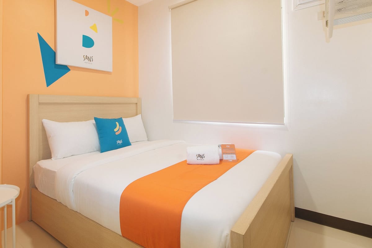 A hotel room with a bed, a bedside table, a window with blinds, and a wall-mounted air conditioning unit.